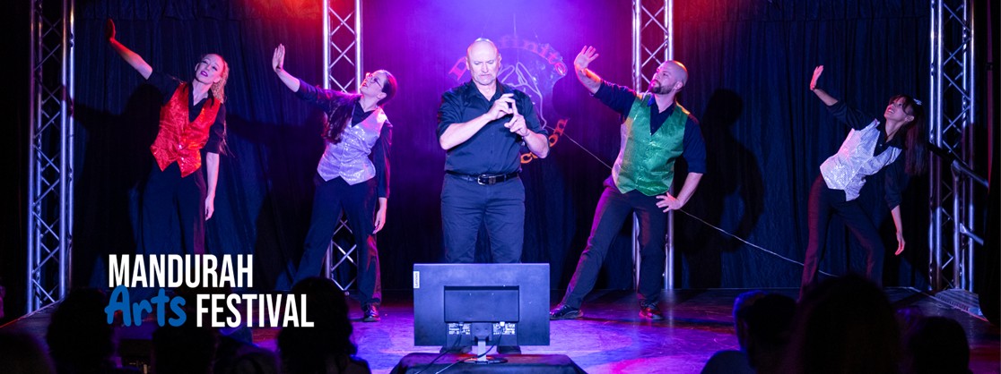 Four people in coloured vests are posed as though dancing, and one man in black holds his hands up, signing a word using Auslan. They are all on a stage lit by purple and red lights